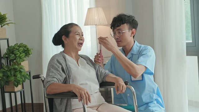 Young male nursing home worker applying makeup to cheerful elderly woman in wheelchair