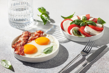 Caprese salad with tomatoes, mozzarella cheese and basil leaves. Eggs, bacon and basil leaves in a plate. Hard light