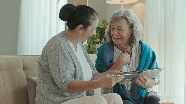 Medium tracking shot of elderly Asian and Caucasian women sitting on couch in living room and discussing pictures in photo album