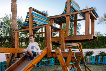 Fototapeta na wymiar A schoolboy boy swinging on swings. Modern stylish eco-style playground made of natural materials. Activities for children on the territory of the hotel Holidays in park