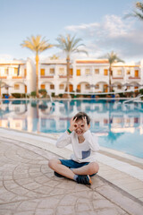 Fototapeta na wymiar A cheerful child in a long-sleeved T-shirt sits by the pool. Rest in the hotel. Happy holidays. Winter or autumn at the resort. Warm evening. swimming pool is heated. Free childhood. child lost a toot
