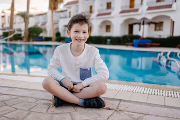 A cheerful child in a long-sleeved T-shirt sits by the pool. Rest in the hotel. Happy holidays. Winter or autumn at the resort. Warm evening. swimming pool is heated. Free childhood. child lost a toot
