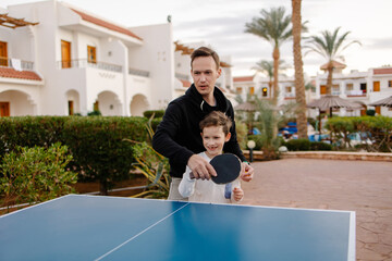 Dad teaches son to play table tennis on vacation at the hotel. The coach and the child are playing ping pong. Sports life active lifestyle. Parenthood fatherhood together. Active outdoor activities. - obrazy, fototapety, plakaty