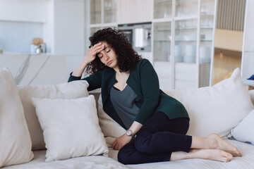 Exhausted curly European woman  sitting on sofa holding her head with eyes closed. beautiful Italian woman resting after a working day at home, headache. sleepy entrepreneur tired after a working week