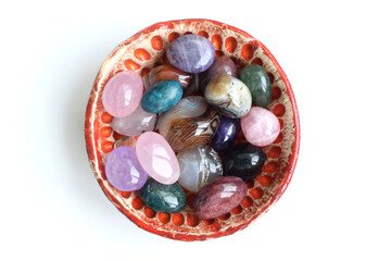 Colorful tumbled semi-precious stones lie in a handmade ceramic bowl. Red bowl with agate, rose quartz, amethyst and apatite on a white background.