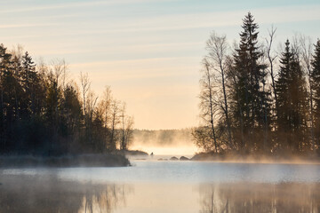 Panoramic view of the forest lake at sunrise. Soft golden sunlight, clear blue sky, reflections on water, fog, frost. Mighty trees. Finland. Nature, seasons, ecology, environment, eco tourism themes