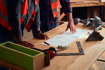 Close-up of men in reflective clothing gesturing at paper near digital tablet in woodworking factory