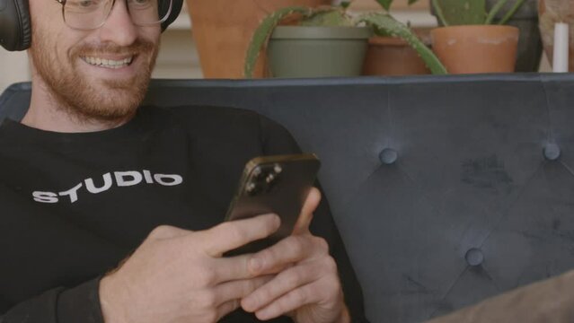 Medium close front angle of a man sitting on a couch scrolling and swiping and typing on an Iphone.