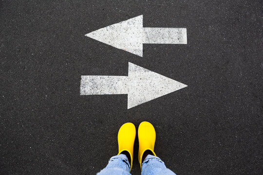 Yellow rubber boots on the asphalt road with drawn arrows pointing to two directions. Making decisions and making choices