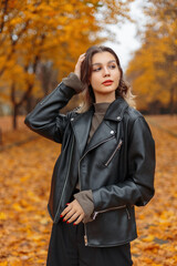 Stylish beautiful young woman with red lips in a fashionable black leather jacket and green sweater walks in a golden park with foliage