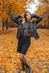Stylish young girl in autumn fashionable clothes with a black leather jacket, a green sweater, shorts, tights and boots walks in an autumn yellow park with golden fall foliage