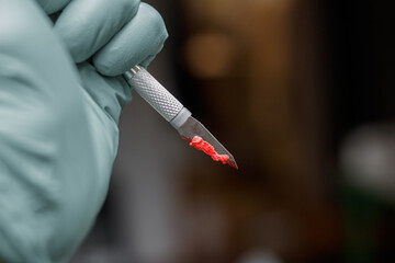 Close up on a gloved hand holding a bloody scalpel covered in organic matter on the left side of the frame. The scalpel is held like a knife ready to stab. Blurred dark background