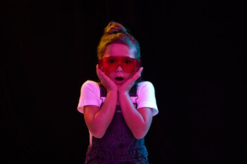 Wow, shock. Fashionable nice-looking little girl looking at camera with surprise isolated over dark background in neon light. Concept of beauty, kids fashion, children emotions