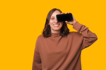 Portrait of young woman holding smartphone over eyes and smiling over yellow background