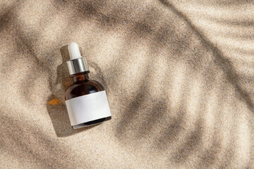 Dropper bottle with blank label on beige sand top view, palm leaf hard shadow. Cosmetic mockup