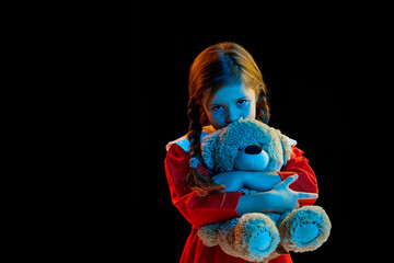 Portrait of beautiful cute little girl with pigtails wearing red festive dress standing with teddy bear isolated over dark background. Concept of children emotions, child psychology