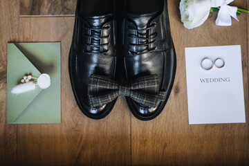 The groom's black shoes, bow tie, boutonniere, gold rings, an envelope with a heart print, a sheet...