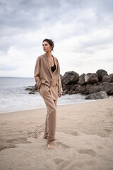 Fantastic  woman walking on the sand beach on a sea background. Beautiful tall model, slim figure. Brunette, beige suit. Fashion, style clothes.