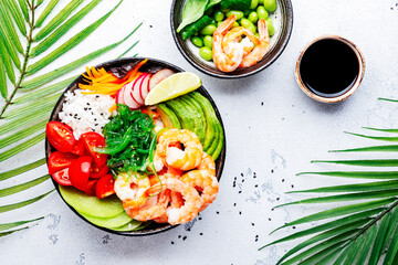 Hawaiian poke bowl with shrimp, avocado, radish, carrot, tomato, chuka seaweed and white rice. Soy sauce, lime and sesame dressing. White table background, palm leaves, top view