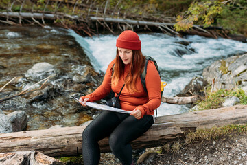 Stylish woman holding paper map with green backpack and relaxing in nature. Travel and wanderlust concept. Amazing chill moment in the forest