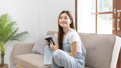 Asian woman sits with a tablet on the sofa in her living room,  Weekend leisure activities, Happiness during vacation at home, Favorite corner of the house, Beautiful woman smiling sweetly.