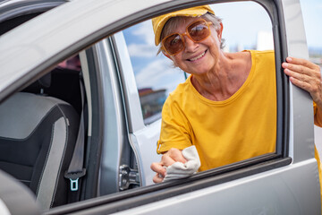 Smiling Caucasian senior woman in yellow t-shirt and hat cleans the car in a self-service car wash...