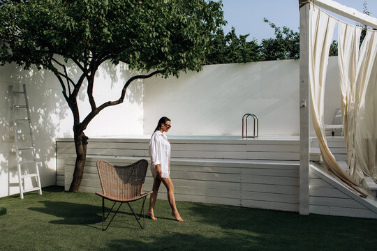 Tanned girl in a white shirt on a naked body. A woman in a white shirt, wearing sunglasses sits on a green grass or lawn. Beach summer vacation, holiday season. Backyard of the house
