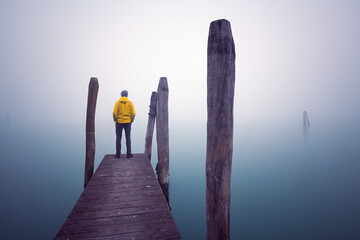 Eine Person steht mit einer knalligen Jacke am Ende eines Bootssteges über dem Wasser und sieht...