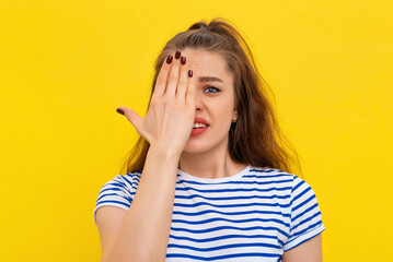 Close up portrait of scared brunette girl, cover half of face and looking at camera with one eye, checking vision, standing in white-blue striped t shirt over yellow background
