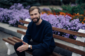 Young happy man rests on bench in park near flowerbed.