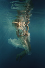 A woman with blond hair in a white dress swims underwater as if in weightlessness