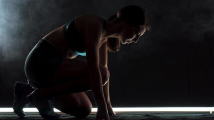 Silhouette female runner in race  start position. Girl in sportswear posing on lit track with smoke in the background.
