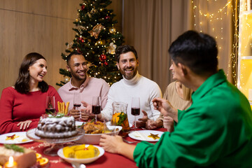 Friends celebrate Christmas eve or New Year holiday paty together sitting at the table. Feast at home group of multi ethnic festive christmas dinner. happy cheerful people clink wine glasses laugh