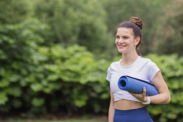 Portrait of happy young caucasian woman exercising yoga outdoors early morning. Beautiful girl with blue yoga mat on green grass in park at summer