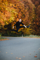 Young attractive man is having fun on road in autumn forest. Guy with short hair wearing in black is jumping in park with yellow leaves trees. Cozy day on countryside