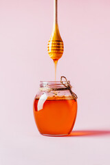 close-up on a jar of honey and a whisk for honey with dripping honey on a pink background