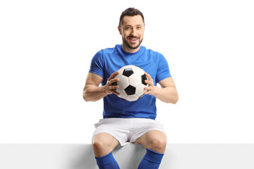 Football player sitting on a blank panel and holding a soccer ball