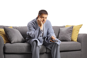 Bored young man in a bathrobe holding a remote control and sitting on a sofa
