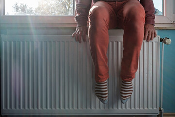 Man warms feet near a slightly warm radiator. Concept of energy crisis and increase in heating costs