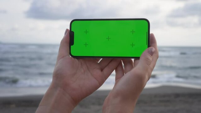 Point of View of woman holds horizontal mobile phone two hands and watching a video on background of ocean beach with cloudy sky. Use green screen for copy space. Chroma key mock-up on smartphone. 