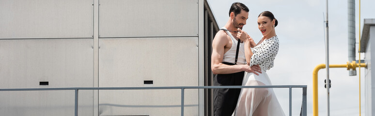 Cheerful and stylish professional dancer touching suspenders on partner on roof, banner