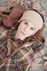 Portrait of a country woman in autumn lying on her back leaning on her bag. Mountain woman with two braids, turban and plaid shirt. Autumn landscape, leaves background.