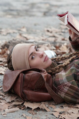 Caucasian country woman reading a book, her head resting on her bag. She wears two braids, a plaid shirt and a bandana for the cold. Autumn landscape with leaves.