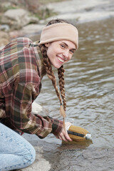 Woman with mountain look in the middle of autumn and filling the canteen to the river and smiling. Countryside caucasian woman with braids, shirt, plaid shirt, bandana. Excursion through the forest. 