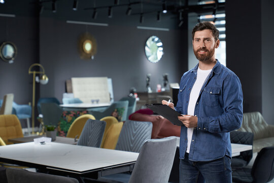 Smiling bearded salesman in denim standing with black tablet in new furniture store. Front view of confident retailer making notes, while looking at camera in expo center. Concept of workplace.