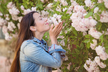 Obraz premium Lifestyle portrait of young stylish woman staying on the street in old town and sniffing the flowers of white roses, wearing blue dress and denim jacket