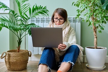 Woman in warm sweater using laptop sitting near heating radiator at home
