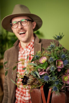 Attractive Laughing Male Person In Eyeglasses, Vintage Clothes And Hat Staying With Flower Bouquet And Briefcase In Flower Store. Looking At Camera. High Quality Vertical Image