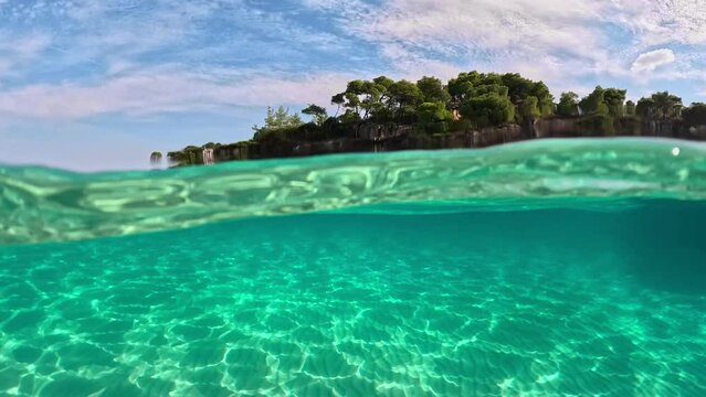 Half underwater slow-motion view of sandy beach bottom and rock with trees. Crystal clear sea water in Sitonia, Halkidiki, Greece. Shot from border of air and water with dome