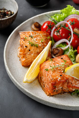 Baked Salmon and green fresh vegetable salad with tomatoes cherry, onion, lemon on ceramic plate. Concept keto diet healthy food. Dinner idea. Close up. Dark background.